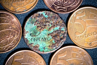 Several coins are displayed on a dark, textured background. Most of the coins have a golden-brown metallic appearance and include embossed text and numbers. One coin, in the center, appears heavily corroded and shows greenish oxidation, contrasting with the others.