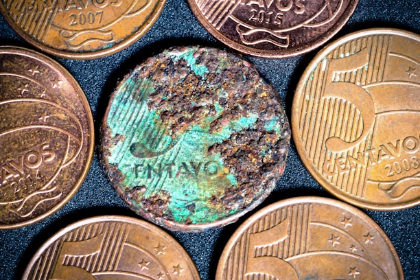 Several coins are displayed on a dark, textured background. Most of the coins have a golden-brown metallic appearance and include embossed text and numbers. One coin, in the center, appears heavily corroded and shows greenish oxidation, contrasting with the others.