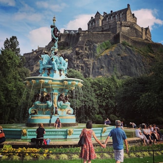 A blue ornate fountain adorned with sculptures, surrounded by tourists, is the central focus. It is set in a lush green park with a rocky cliff and a large, historic castle prominently in the background. People are sitting and standing around, enjoying the scenery.