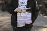 A person holds a clapperboard used in film production. The clapperboard includes details such as scene number, take number, and the name Hunter Moranville. The person is wearing a white T-shirt with text and graphics related to filmmaking and a black jacket.