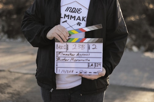 A person holds a clapperboard used in film production. The clapperboard includes details such as scene number, take number, and the name Hunter Moranville. The person is wearing a white T-shirt with text and graphics related to filmmaking and a black jacket.