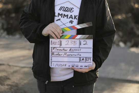 A person holds a clapperboard used in film production. The clapperboard includes details such as scene number, take number, and the name Hunter Moranville. The person is wearing a white T-shirt with text and graphics related to filmmaking and a black jacket.