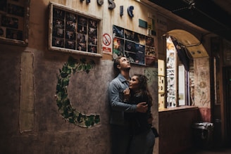 A couple embraces in a warmly lit urban setting, surrounded by posters and photographs on old, textured walls. The environment has a rustic and artistic feel, with a prominent 'no smoking' sign adding to the eclectic decor.