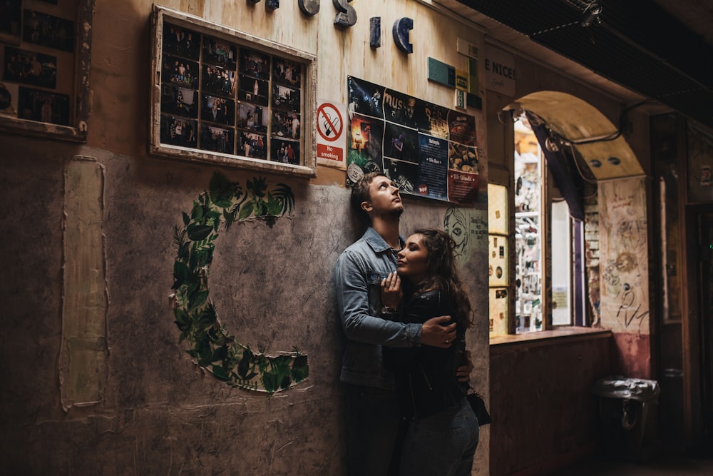 man in blue chambray long-sleeved shirt embracing woman in black jacket and both leaning on wall