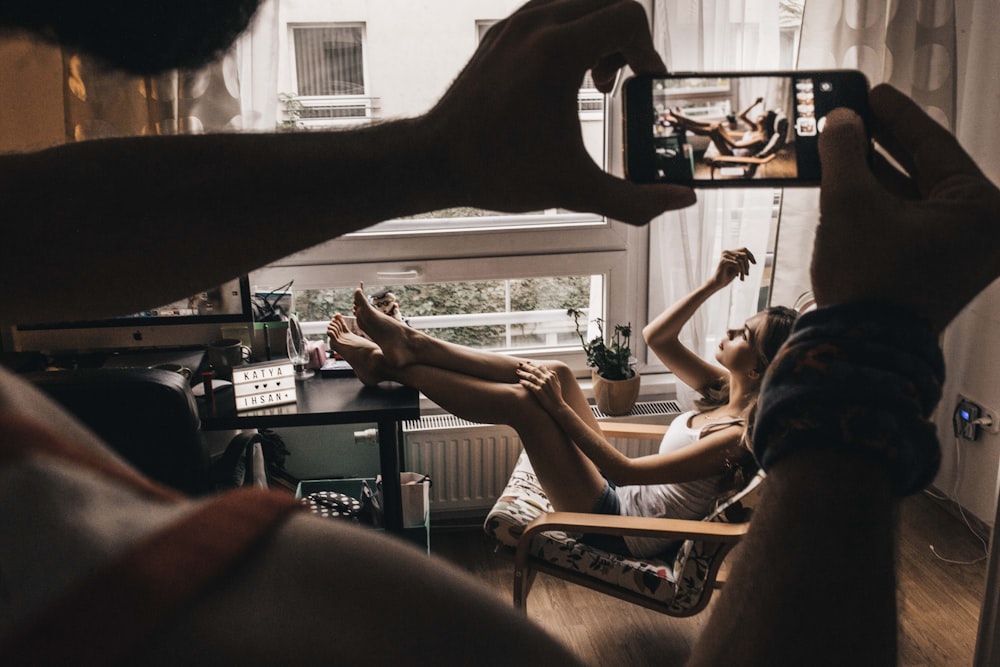 man taking photo of woman sits on chair near window using smartphone