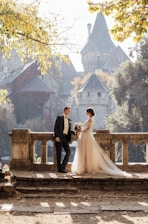 A couple dressed in wedding attire is standing on an old stone balcony or terrace, surrounded by lush greenery and historic architecture. The man is wearing a black suit while the woman is in a long, flowing white wedding dress with a bouquet of flowers. The background includes an old castle with pointed towers and a serene atmosphere. Sunlight filters through the trees, creating a warm and romantic ambiance.
