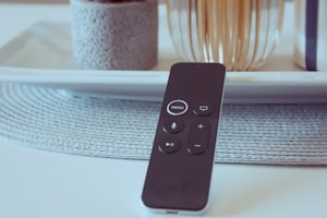 A sleek, black remote control lies on a textured gray placemat with a modern, minimalistic setting in the background. The remote features buttons including menu, volume, and play controls. Behind the remote, there is a decorative arrangement with a stone-like vase and fluted glass or ceramic objects, all placed on a clean, white tray.