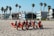 A group of women wearing red swimsuits and pink visors sitting on beach chairs set on a sandy beach. They are facing towards the camera with some looking away. Behind them, there are tall palm trees and modern buildings. The sky is clear and blue, and there are a few people walking in the background.