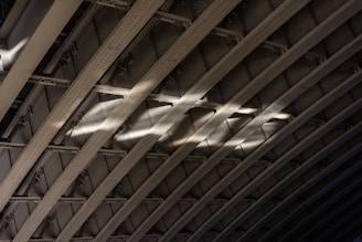 Curved metal beams create a pattern of arches with light casting across them, highlighting the texture and structure. The rivets and shadows accentuate the industrial design.