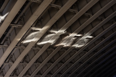 Curved metal beams create a pattern of arches with light casting across them, highlighting the texture and structure. The rivets and shadows accentuate the industrial design.