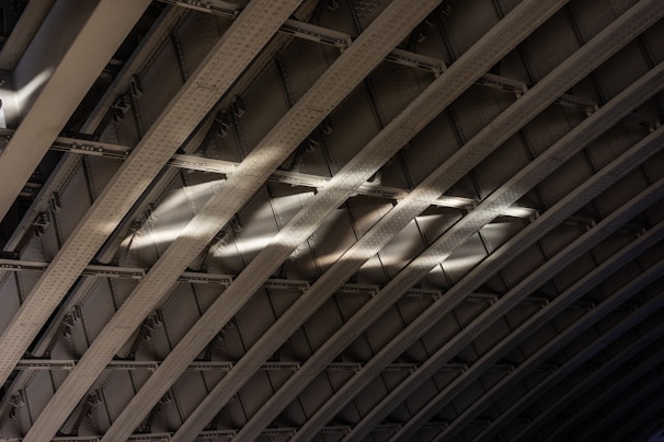 Curved metal beams create a pattern of arches with light casting across them, highlighting the texture and structure. The rivets and shadows accentuate the industrial design.