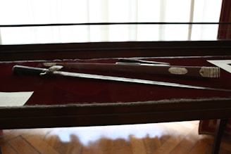 A sword and its sheath are displayed in a glass case with a dark red background. The sword has an ornate handle with intricate detailing. There is text on plaques next to the items.