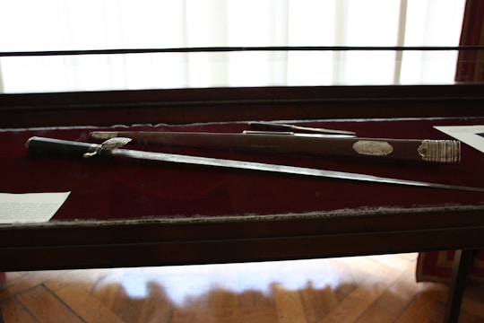 A sword and its sheath are displayed in a glass case with a dark red background. The sword has an ornate handle with intricate detailing. There is text on plaques next to the items.