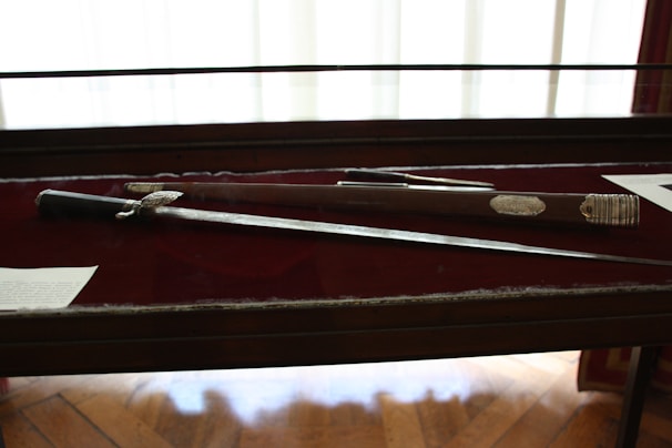 A sword and its sheath are displayed in a glass case with a dark red background. The sword has an ornate handle with intricate detailing. There is text on plaques next to the items.