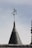 A conical roof topped with a weather vane displaying a flag. The surface of the roof is covered with dark shingles, and the sky in the background is clear with a light greyish hue.
