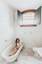 A pregnant woman is relaxing in a vintage-style bathtub located in a white-tiled bathroom. The room features a large window with wooden shutters partially open, allowing natural light to enter. The floor is tiled, and there’s a roll of toilet paper on a holder fixed to the wall near the bathtub.