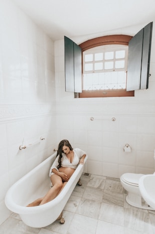 A pregnant woman is relaxing in a vintage-style bathtub located in a white-tiled bathroom. The room features a large window with wooden shutters partially open, allowing natural light to enter. The floor is tiled, and there’s a roll of toilet paper on a holder fixed to the wall near the bathtub.