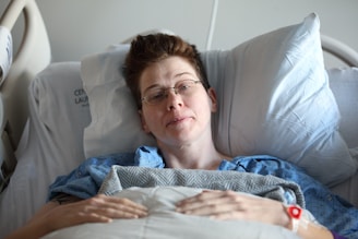 A person wearing glasses is lying in a hospital bed, covered with a blanket. They appear to be awake and resting, wearing a hospital gown with a medical wristband visible on their wrist.