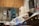 A person with short hair rests in a hospital bed with an oxygen tube connected to their nose. The room contains medical equipment, including a monitor and various devices mounted on the wall. There are wooden cabinets and a green bag hanging in the background.