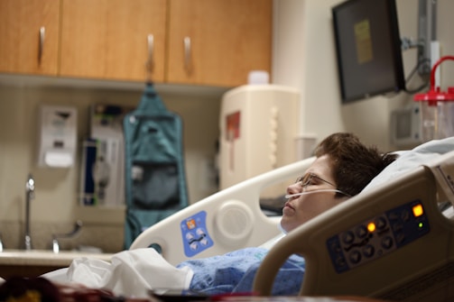 A person with short hair rests in a hospital bed with an oxygen tube connected to their nose. The room contains medical equipment, including a monitor and various devices mounted on the wall. There are wooden cabinets and a green bag hanging in the background.