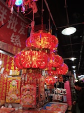 A vibrant market stall filled with red and gold Chinese lanterns, each intricately adorned with traditional patterns and symbols. The stall also displays a variety of festive decorations, including red banners with golden Chinese characters, and posters featuring traditional imagery. The ambient lighting is warm, predominantly red, creating a lively and festive atmosphere, typical of preparations for a celebration such as the Lunar New Year.