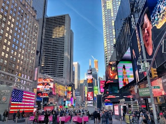 A bustling cityscape with towering skyscrapers and vibrant electronic billboards. People are walking on the busy street, surrounded by colorful advertisements. The area is lively and energetic, with a clear blue sky above.