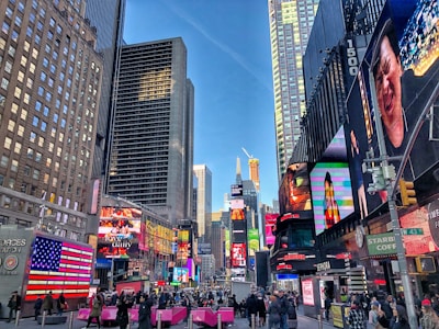 A bustling cityscape with towering skyscrapers and vibrant electronic billboards. People are walking on the busy street, surrounded by colorful advertisements. The area is lively and energetic, with a clear blue sky above.