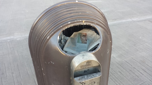 A metallic trash can with a newspaper partially protruding from the opening. The newspaper displays a partially visible image of a face.