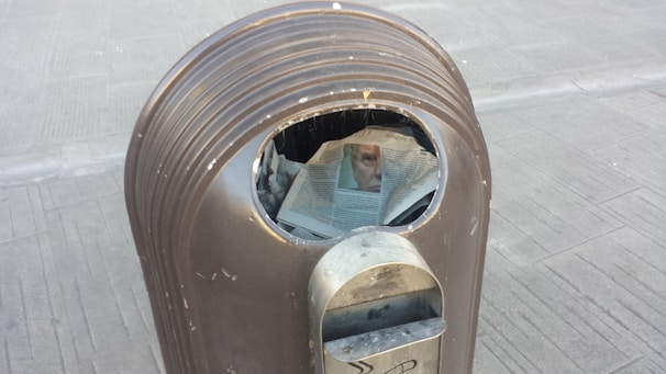 A metallic trash can with a newspaper partially protruding from the opening. The newspaper displays a partially visible image of a face.