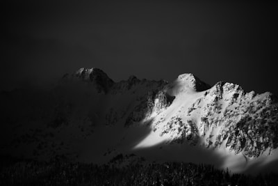 A dramatic black and white photograph of a mountain range with snow-covered peaks. The sunlight casts shadows across the rugged landscape, highlighting the textures of the rocks and snow. The foreground is a dark silhouette of a forest, creating a stark contrast with the illuminated mountains.