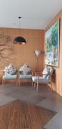 A cozy and modern interior space featuring two light blue armchairs adorned with patterned cushions. A stylish pendant lamp hangs from the ceiling, and a unique branch-like wall decor adds an artistic touch. A large painting depicting a vibrant outdoor scene is mounted on the wooden-paneled wall. A sleek floor lamp with a tripod base stands beside the chairs.