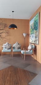 A cozy and modern interior space featuring two light blue armchairs adorned with patterned cushions. A stylish pendant lamp hangs from the ceiling, and a unique branch-like wall decor adds an artistic touch. A large painting depicting a vibrant outdoor scene is mounted on the wooden-paneled wall. A sleek floor lamp with a tripod base stands beside the chairs.