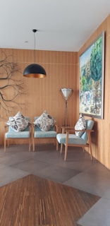 A cozy and modern interior space featuring two light blue armchairs adorned with patterned cushions. A stylish pendant lamp hangs from the ceiling, and a unique branch-like wall decor adds an artistic touch. A large painting depicting a vibrant outdoor scene is mounted on the wooden-paneled wall. A sleek floor lamp with a tripod base stands beside the chairs.