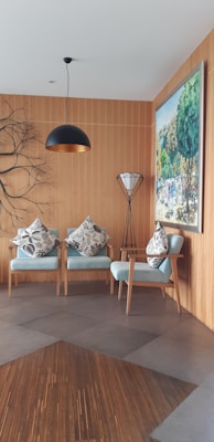 A cozy and modern interior space featuring two light blue armchairs adorned with patterned cushions. A stylish pendant lamp hangs from the ceiling, and a unique branch-like wall decor adds an artistic touch. A large painting depicting a vibrant outdoor scene is mounted on the wooden-paneled wall. A sleek floor lamp with a tripod base stands beside the chairs.