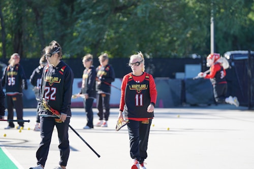 A group of female athletes wearing black and red sports uniforms with Maryland written on them are standing outdoors on a court. They are holding lacrosse sticks and appear to be involved in a training session or preparing for a game. The background includes green foliage and some equipment.