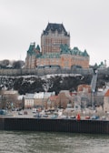 A grand castle-like hotel with turrets and intricate architecture sits atop a hill. Below, a quaint town with colorful, European-style buildings lines the waterfront, while cars are parked along the road. The scene is tranquil, with overcast skies and a calm river in the foreground.
