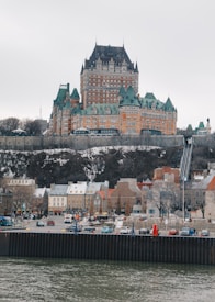 A grand castle-like hotel with turrets and intricate architecture sits atop a hill. Below, a quaint town with colorful, European-style buildings lines the waterfront, while cars are parked along the road. The scene is tranquil, with overcast skies and a calm river in the foreground.
