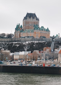 A grand castle-like hotel with turrets and intricate architecture sits atop a hill. Below, a quaint town with colorful, European-style buildings lines the waterfront, while cars are parked along the road. The scene is tranquil, with overcast skies and a calm river in the foreground.