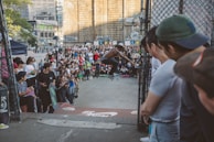 A skateboarder is performing a trick in the middle of an urban setting, surrounded by an audience watching closely. The scene is lively with a diverse crowd gathered, some holding skateboards. Buildings and graffiti are visible in the background, contributing to the urban atmosphere.
