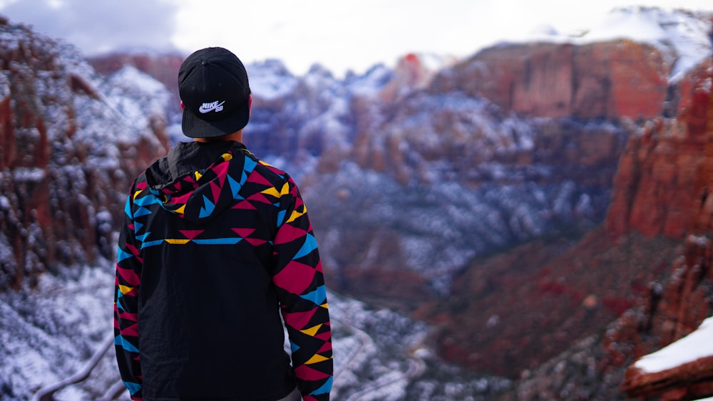 man wearing black, blue, and yellow hoodie and fitted cap facing snow covered mountain