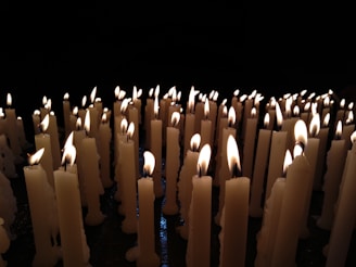 Rows of white candles are lit, with bright flames flickering in a dark setting. The glow from the candlelight casts a warm, serene illumination, highlighting the candle wax textures.