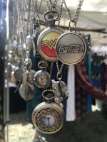 A collection of vintage-style pocket watches and pendant necklaces are hanging on display. The pocket watches feature intricate designs and logos, including a red and yellow insignia, a famous soft drink logo, and floral engravings. The soft lighting and outdoor setting suggest a market or fair environment.