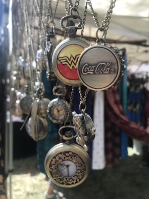A collection of vintage-style pocket watches and pendant necklaces are hanging on display. The pocket watches feature intricate designs and logos, including a red and yellow insignia, a famous soft drink logo, and floral engravings. The soft lighting and outdoor setting suggest a market or fair environment.
