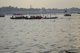 Boats with people in life jackets are floating on a wide river, surrounded by many birds flying above the water. The background shows a distant shoreline with a silhouette of trees and some structures. The sky is overcast, contributing to an overall tranquil atmosphere.