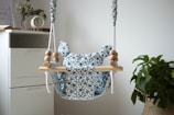 A baby swing with a floral patterned fabric is suspended with ropes and wooden beads. It is situated near a white drawer, and a small clock and a book are on top of the drawer. A plant with large leaves is in a woven basket beside the swing.