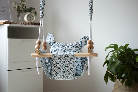 A baby swing with a floral patterned fabric is suspended with ropes and wooden beads. It is situated near a white drawer, and a small clock and a book are on top of the drawer. A plant with large leaves is in a woven basket beside the swing.