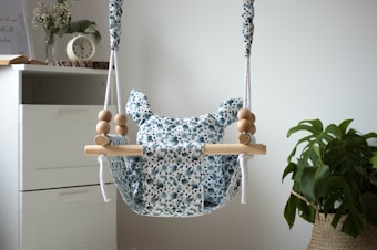 A baby swing with a floral patterned fabric is suspended with ropes and wooden beads. It is situated near a white drawer, and a small clock and a book are on top of the drawer. A plant with large leaves is in a woven basket beside the swing.