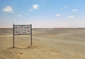 A sparse desert landscape with a directional signpost indicating camping facilities 1500 meters ahead. The sign reads 'Camping Secret de Sahara' and mentions a café and restaurant. The horizon features distant sandy dunes under a clear blue sky with a few scattered clouds.