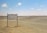 A sparse desert landscape with a directional signpost indicating camping facilities 1500 meters ahead. The sign reads 'Camping Secret de Sahara' and mentions a café and restaurant. The horizon features distant sandy dunes under a clear blue sky with a few scattered clouds.