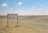 A sparse desert landscape with a directional signpost indicating camping facilities 1500 meters ahead. The sign reads 'Camping Secret de Sahara' and mentions a café and restaurant. The horizon features distant sandy dunes under a clear blue sky with a few scattered clouds.